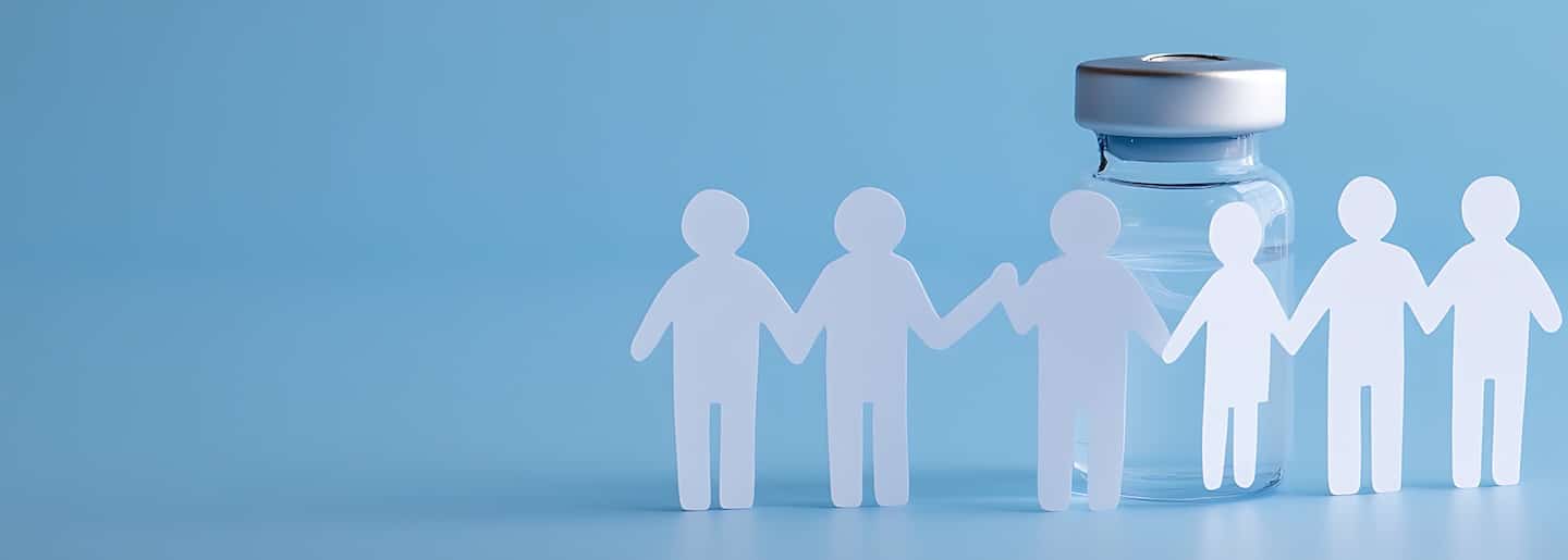 A line of paper dolls holding hands in front of a vaccine vial on a light blue surface symbolizing health and community immunity in a medical context.